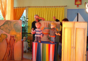 Elementy scenografii – rysunek drzewa, szafy oraz kanapy. Przy rysunku kanapy umieszczonym w centralnej części animatorzy trzymają dwie sporej wielkości lalki przedstawiające dwóch chłopców.